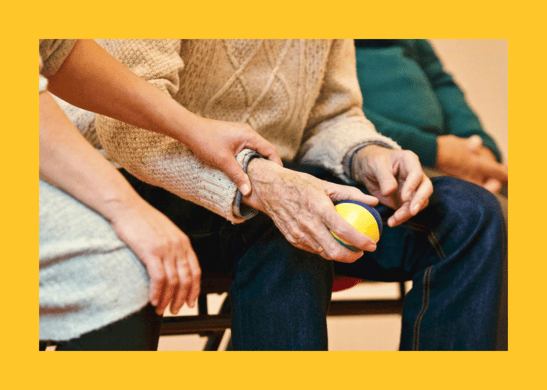 A photo in in a yellow frame of a younger adult lending a helping hand to an older adult who is participating in an activity with a ball.